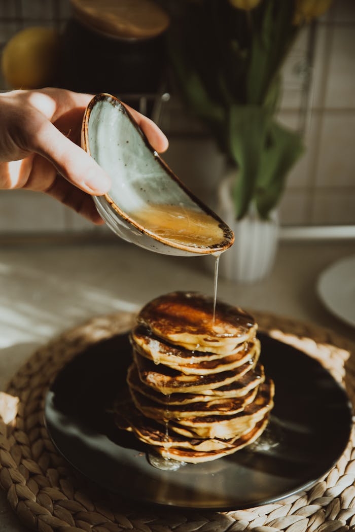 A hand pours honey on a stack of pancakes in a sunlit kitchen, creating a warm, inviting breakfast scene.